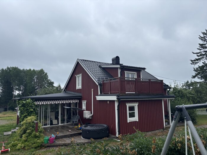 Rött hus med grått tak, utbyggnad ovanför balkongen diskuteras för sovrum. Veranda med växter och lekredskap i förgrunden, molnig himmel.