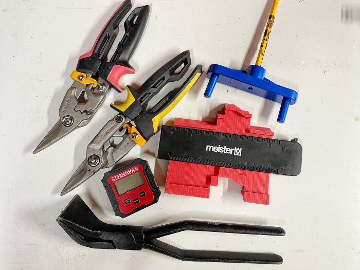 Verktyg inklusive två plåtsaxar, en profilmall, en centrummsrkör, en vinkelmätare och en falstång liggande på en vit yta.