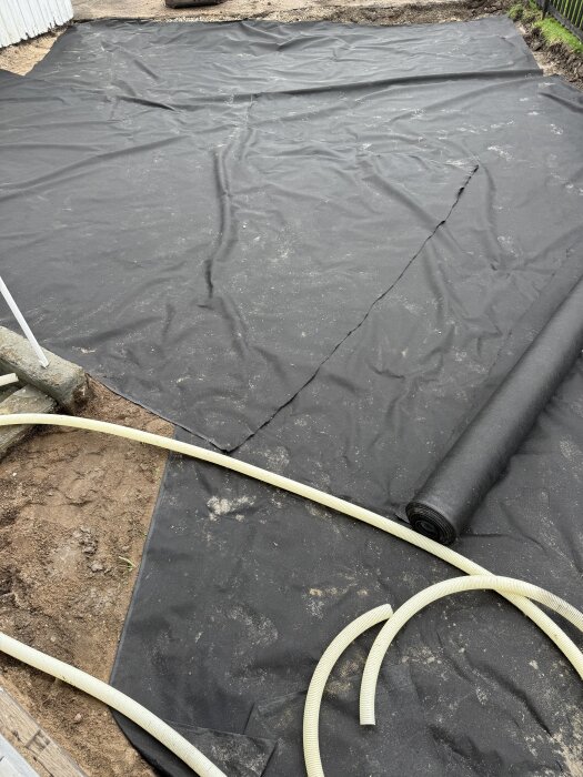 Markduk utrullad över mark med dräneringsslang ovanpå, rullar av duk synliga. Förberedelse för altanprojekt med dränering till avlopp.