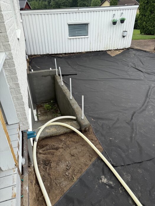 Dräneringsslang och markduk installerad vid hus, förbereder för grustäckning för att lösa vattenansamling vid altanhörn.
