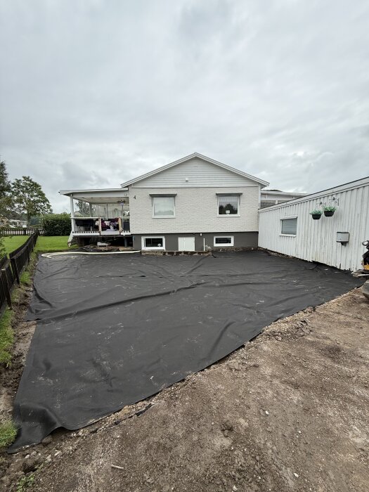 Markduk utrullad över en gårdsplan vid ett hus, förberedd för dräneringslösning med slang för att leda bort vatten vid hörnet av altanen.