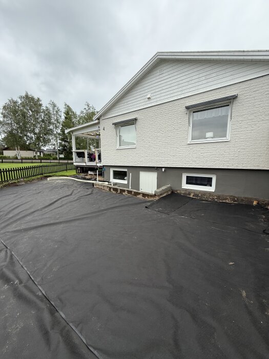 Markduk utrullad över marken vid ett hus med altan, förberett för dränering med slang för att hantera regnvatten och undvika vattenansamlingar.