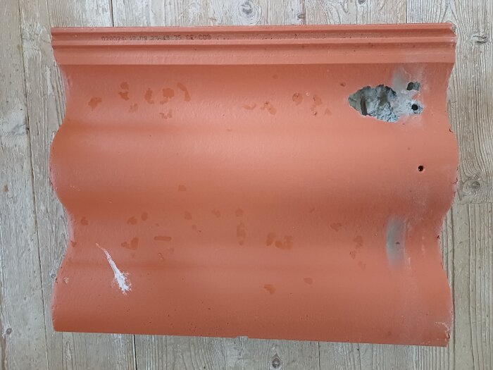 Trasig takpanna med hål och söndervittring på höger sida, liggande på ett trägolv.