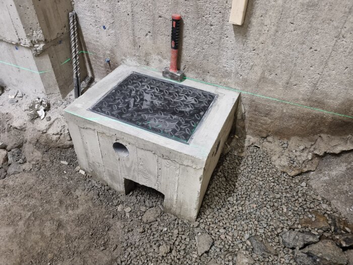Betonglåda med monterad lucka i metall och ett verktyg ovanpå, omgiven av sten och grus; en del av ett källarprojekt.