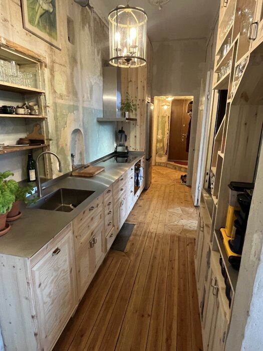 Renoverat kök med träpaneler, diskbänk av rostfritt stål, öppen hylla, och hängande taklampa. Trägolv och modern design.