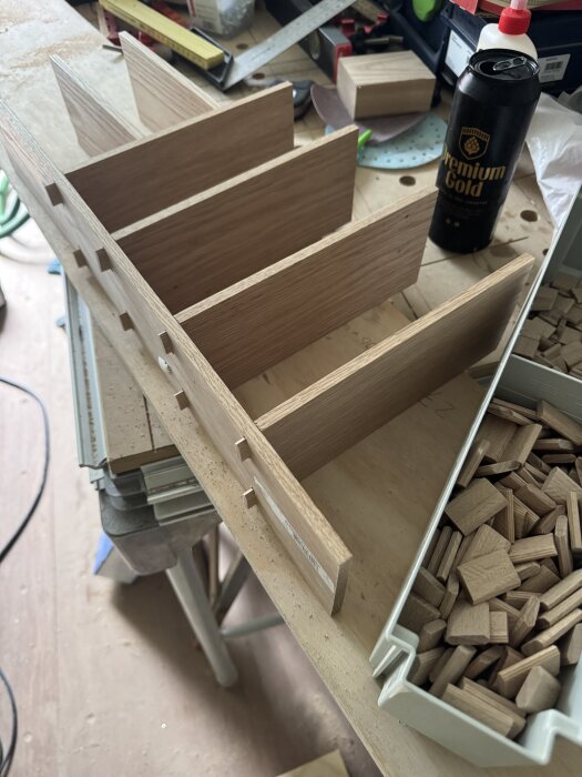 Trälåda under konstruktion med dominobrickor och verktyg på arbetsbänk. Kapskivor och en burk syns bredvid.