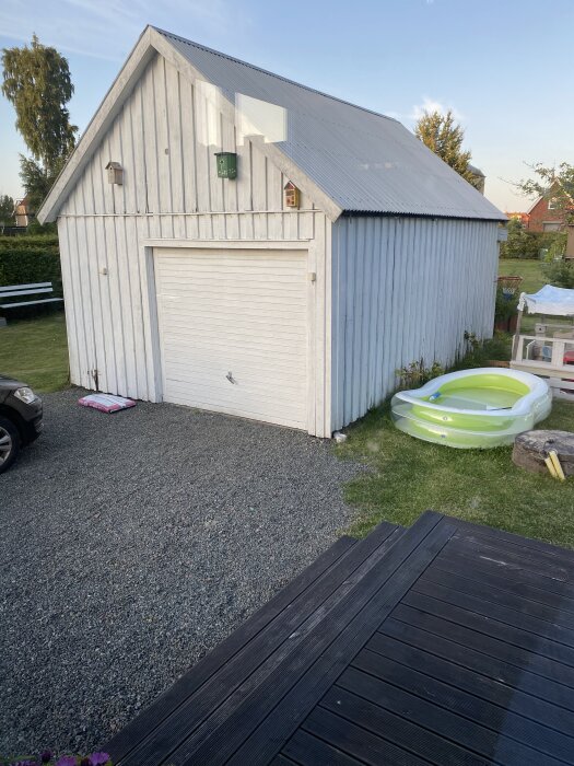 Ett gammalt vitt garage med plåttak och stängd port, beläget vid en grusad uppfart bredvid en uppblåsbar pool och trädäck.