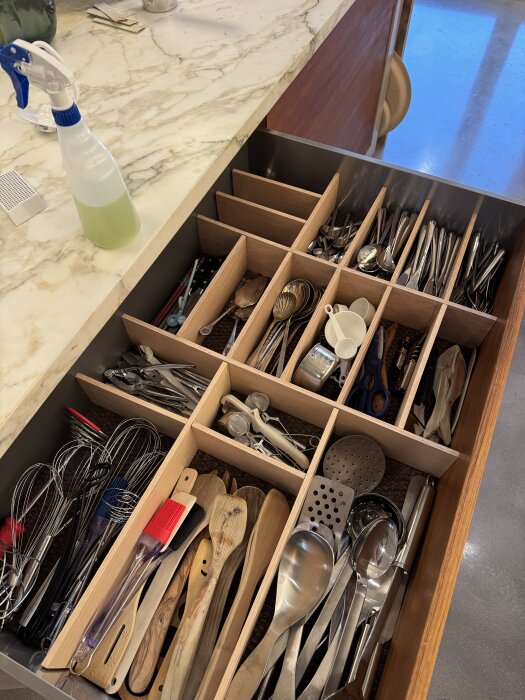 Besticklåda med träavdelare organiserar vispar, slevar, bestick och köksredskap i en kökslåda med marmorbänkskiva och rengöringsspray.