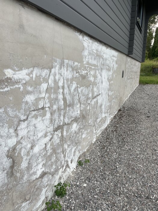 Husfasad med fläckvis puts och grå panel ovanför, bredvid en grusgång.