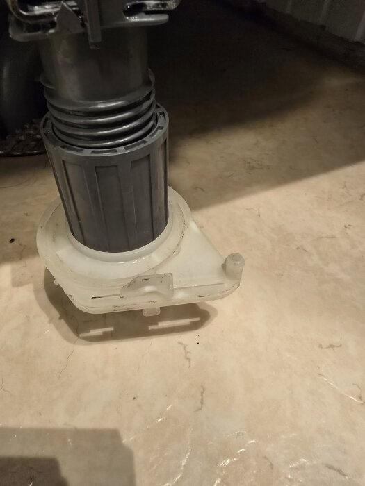 En grå cylinder med plastdelar som inte sitter fast ordentligt, på golv, med en pigg synlig utan tydlig funktion.