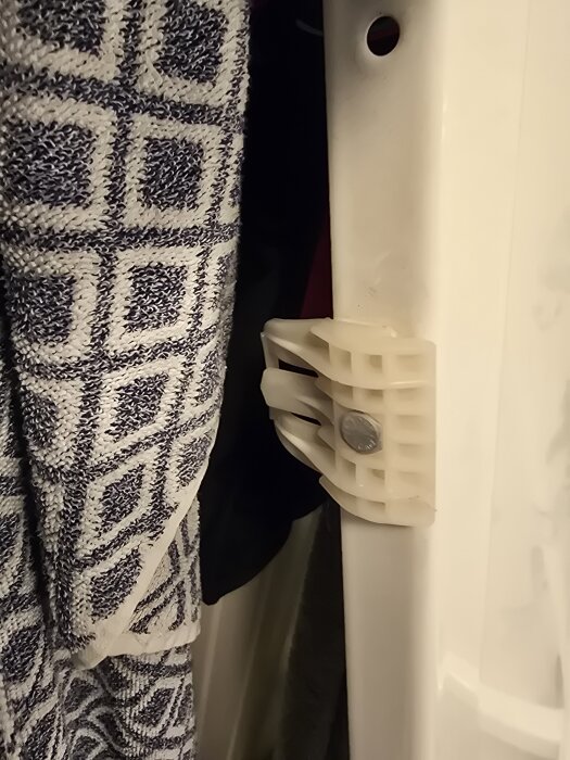 Vit plastkomponent vid kanten av ett badkar med pigg och hål, bredvid en mönstrad handduk.