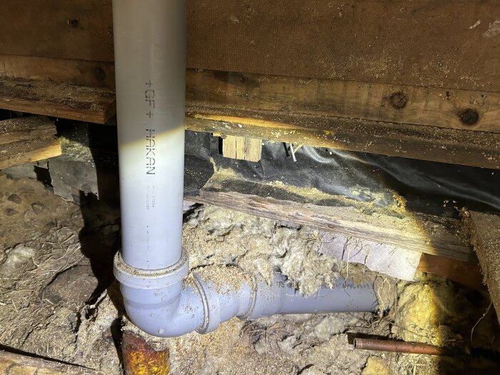 Avloppsrör med 90-gradersböj under golv i stuga, omgivet av isoleringsmaterial och träbjälkar.