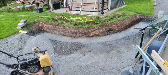 Markerat område för murbygge bredvid friggebod, verktyg och makadam synliga, gräsmatta i bakgrunden.