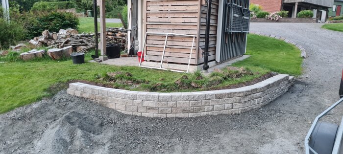 En mur av Benders Stackstone vid en friggebod, omgiven av grönska och en grusgång, med träförvaring och en tom ram bredvid boden.
