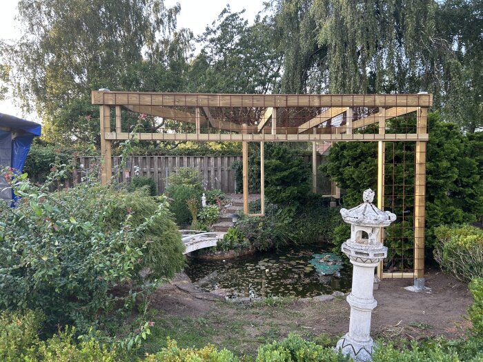 Ett pergola-bygge i en trädgård med en damm och en stenlykta i förgrunden, omgiven av träd och växter.