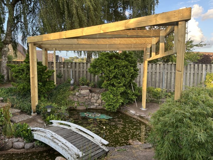 En träpergola med gröna växter runt omkring och en liten bro över en damm i en trädgård.