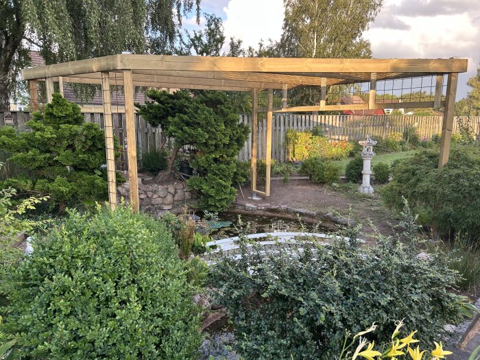 Trästomme för pergola i trädgård under uppbyggnad, omgiven av buskar och en liten damm, med fontän och stenlyktor i bakgrunden.