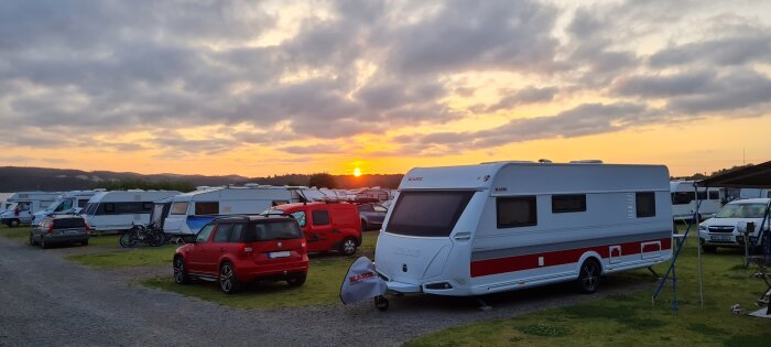 Campingplats i gryningen med flera husvagnar och bilar, där solen håller på att gå upp bakom träden.