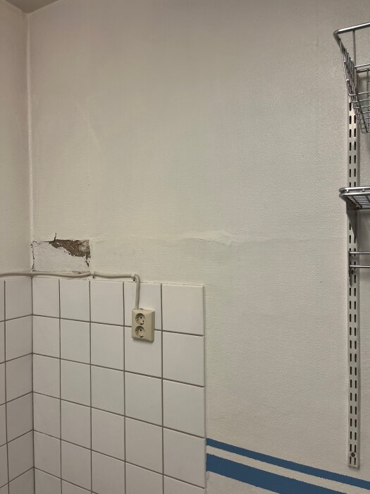 Vägg i tvättstuga med vita kakelplattor, trasig vävtapet och eluttag. Synlig skada där tapeten är borttagen.