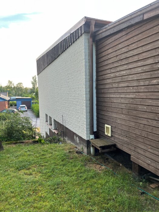 Vitt hus med suterräng, anslutet uterum i trä, stuprör åt sidan, grönskande tomt, buskar och parkerade bilar i bakgrunden.