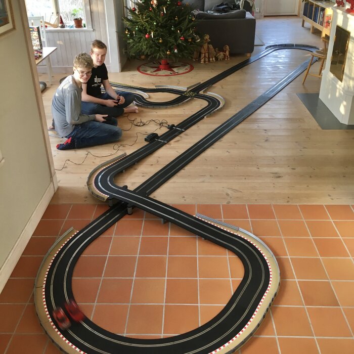 Två personer leker med en bilbana på ett trägolv bredvid en julgran. Golvet är lutat och oljat och visar tecken på långvarig användning.