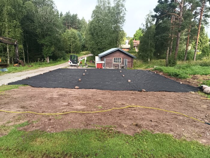 Svart duk lagd på marken framför ett litet trähus med grävskopa och träd i bakgrunden.