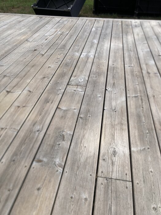 Trädäck av åldrat trä med synliga kvistar och ljusa partier, omgiven av en gräsmatta och möbler i bakgrunden.