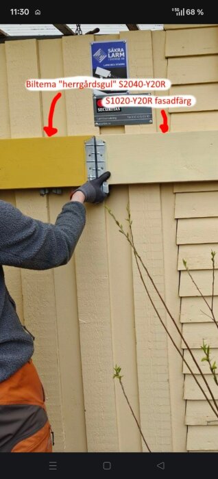 Person monterar en gul träbräda på en gulmålad yttervägg med hjälp av ett gångjärn. Säkerhetsskyltar syns ovanför.