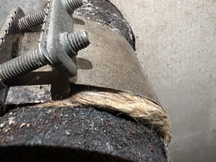 Närbild av rostigt metallrör med fäste och isolerande material runtom, potentiellt asbesthaltiga brandsäkra skivor och ull, del av kamininstallation.