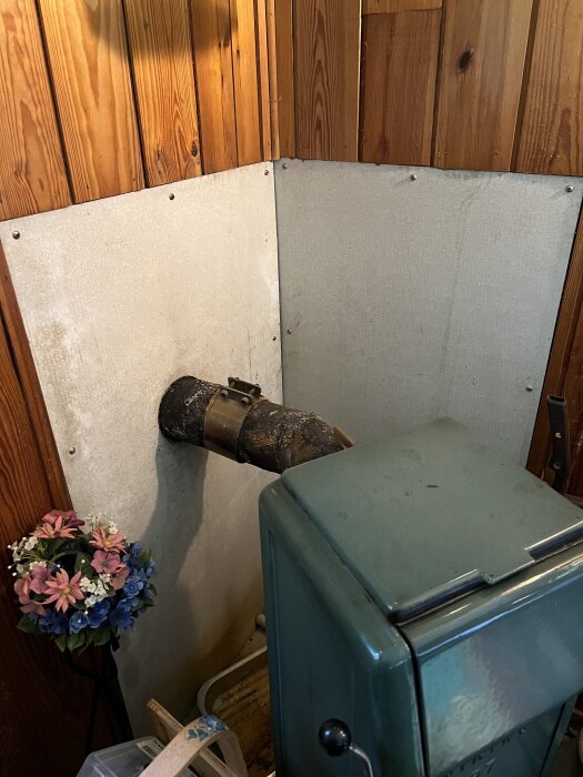 Gammaldags kamin med anslutet rostigt metallrör inomhus, omgiven av träpanel och vit brandsäker skiva. Blommor står bredvid kaminen.