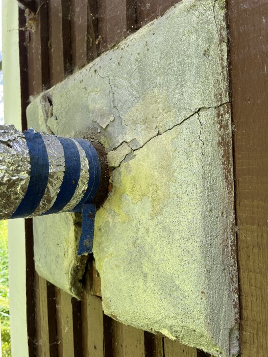 Rostigt metallrör med blå tejp, anslutet till en vägg av sprucken brandsäker skiva på ett trähus; potentiell asbestfara vid renovering.