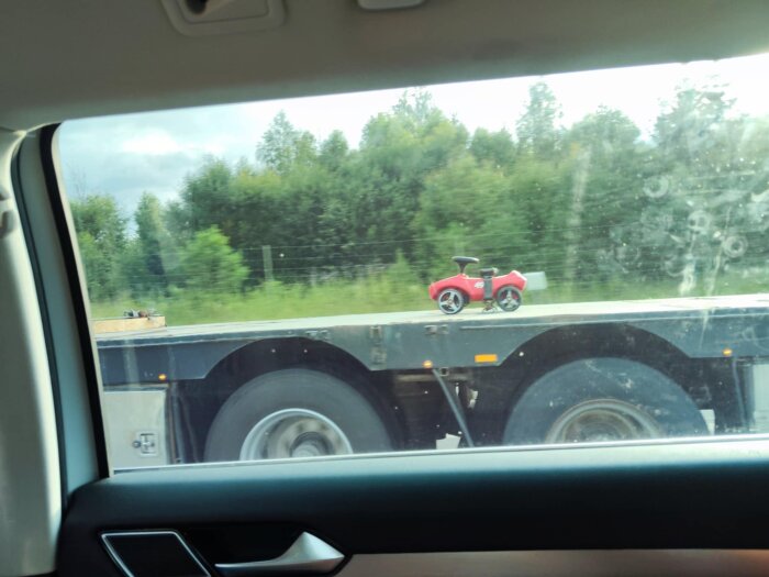 Litet rött leksaksfordon fastsatt med spännband på en stor lastbilsflak, sett från bil.