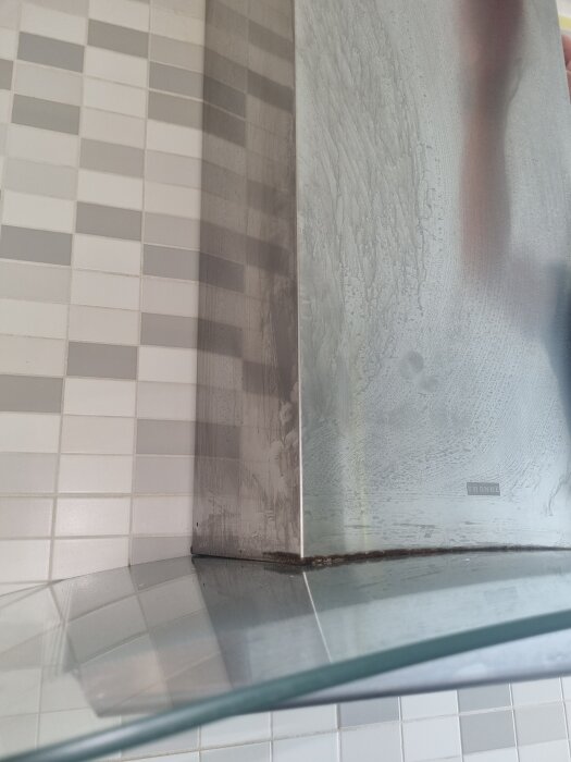 Fläckig yta på rostfri köksfläkt, märkt "Franke", med smuts runt kanterna. Reflektion i glashylla och kakelvägg i bakgrunden.