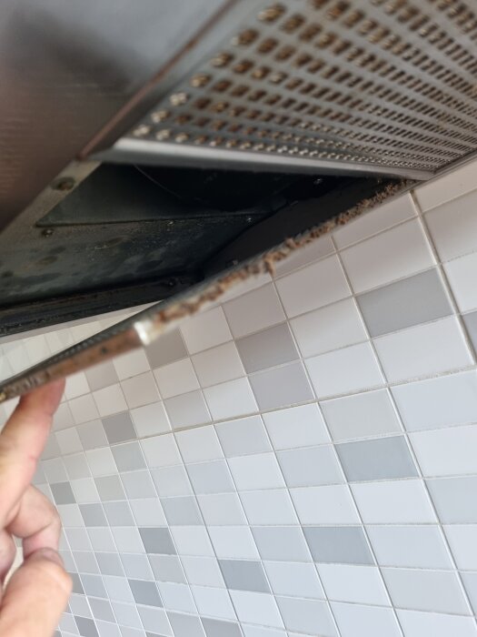 Utsugfläkt under köksskåp med smutsspår i hörnet, nära kaklad vägg. Finger öppnar fläktfilter.