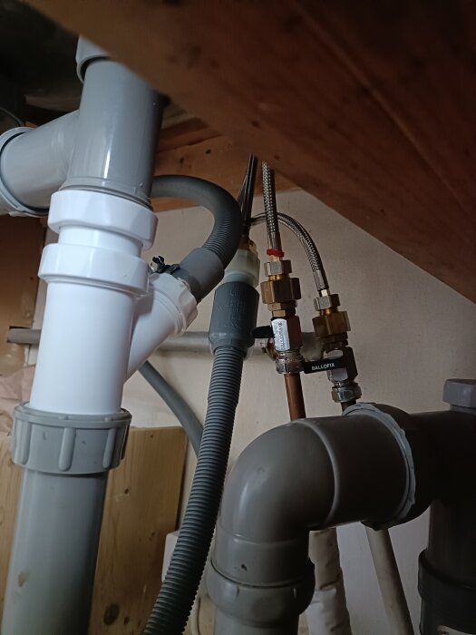 Rör och slangar under diskbänk, del av diskmaskininstallation. Grå och vita plastkomponenter, kopplade till vattenlås och metallventiler.