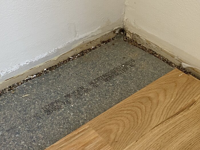 Underlag av grus och träplankor med plast i ett vardagsrum redo för renovering, hörn där en del av plasten är synlig.