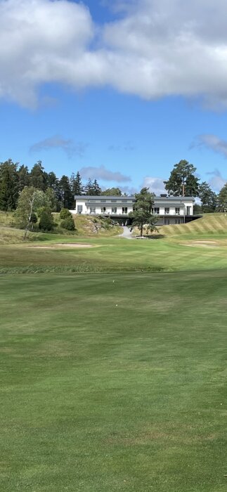 Golfbana med gröna fairways, vit byggnad i bakgrunden, blå himmel och moln.