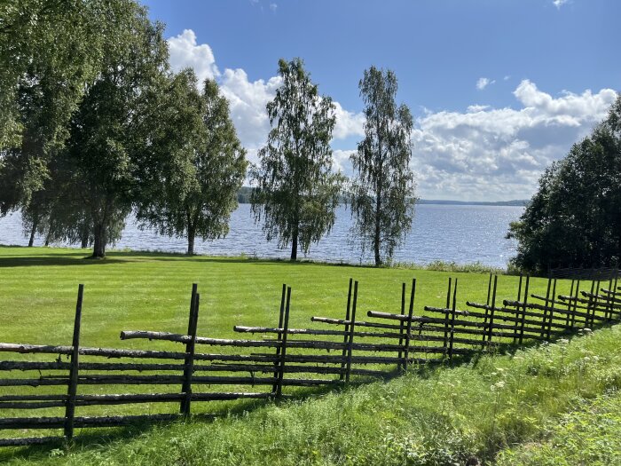 Grön gräsmatta med trästaket i förgrunden, träd i bakgrunden och en sjö under en solig himmel med moln.