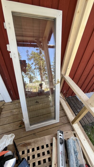 Gammalt fönster lutat mot röd husvägg med utsikt över skog, omgiven av verktyg och byggmaterial på träaltan.