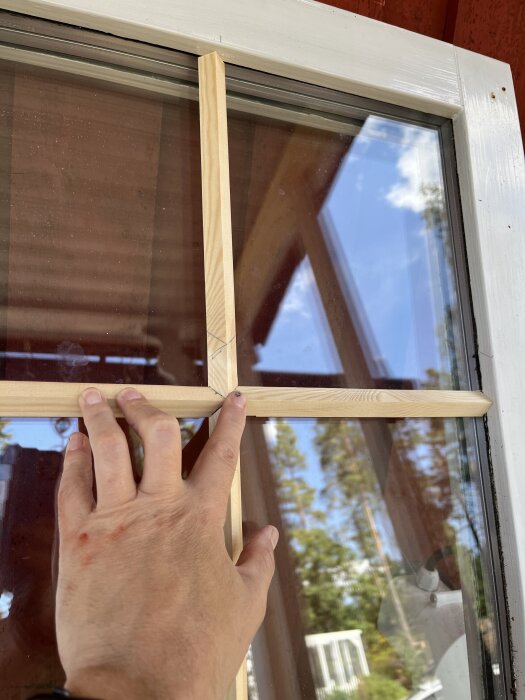 Hand håller trälist mot fönster för att skapa spröjs, blå himmel och träd i bakgrunden.