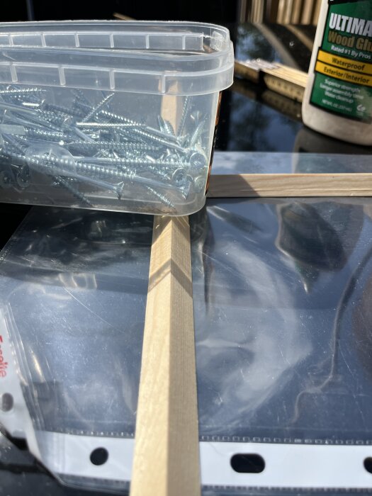 Behållare med skruvar, trälist och flaska med trälim på ett bord i solljus.