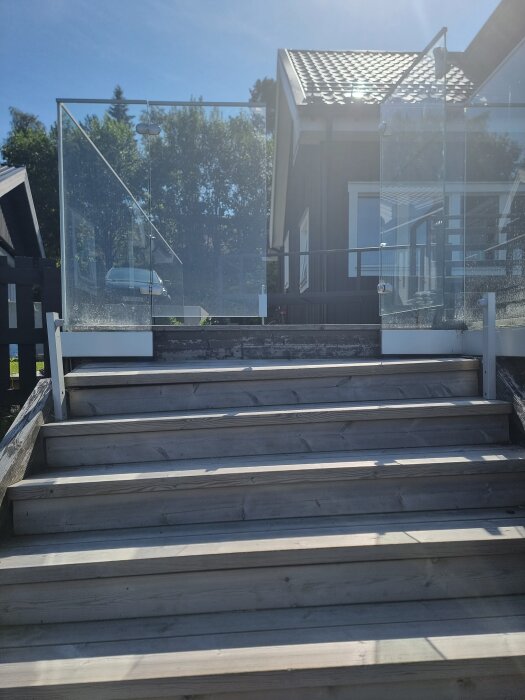 Trappa av trä som leder ned från altan, med glasräcke. Trappan är sned och har en betydande lutning. Hus och träd i bakgrunden.