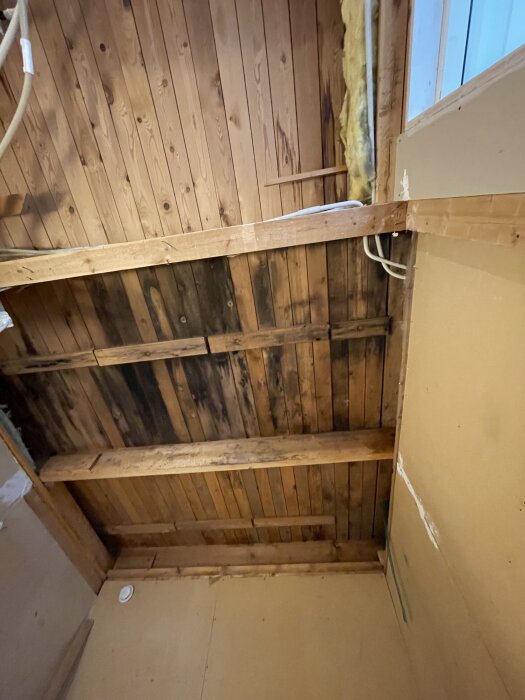 Trätak i förråd med tydlig missfärgning från tidigare vattenläcka, omgiven av isolering och träbalkar.