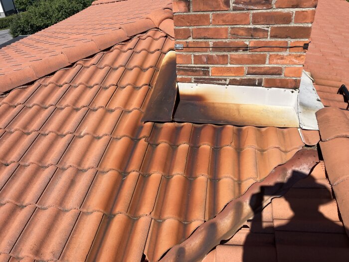 Murad skorsten omgiven av taktegel och delvis rostigt plåtarbete. Färgflagning syns på det falsade taket runt skorstenen.