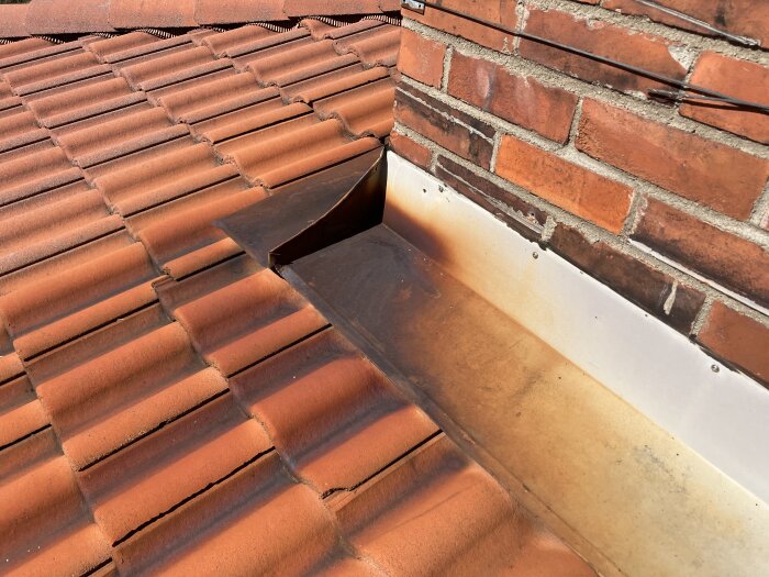 Plåtdetalj vid röd tegeltak och murad skorsten, med synlig ytrost på metallytan.