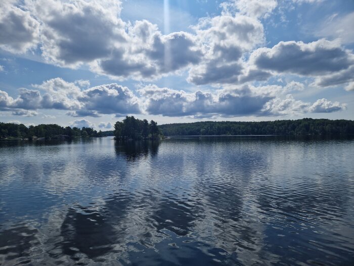 Sjölandskap med spegelblankt vatten, molnig himmel, och skogsklädd horisont i bakgrunden.