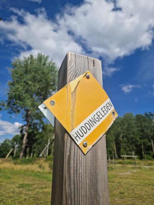 Gul skylt med texten "Huddingeleden" på en trästolpe mot en bakgrund av träd och blå himmel med moln.