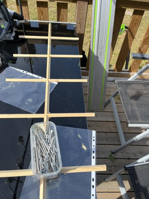 En arbetsbänk med skruvar i en plastlåda, träpinnar, ett måttverktyg och en sänksåg, med en altan i bakgrunden.