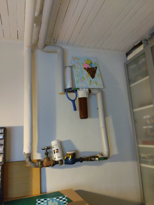 Rörinstallation med ventiler och vattenmätare på en vägg, dekorerad med färgglad tavla.