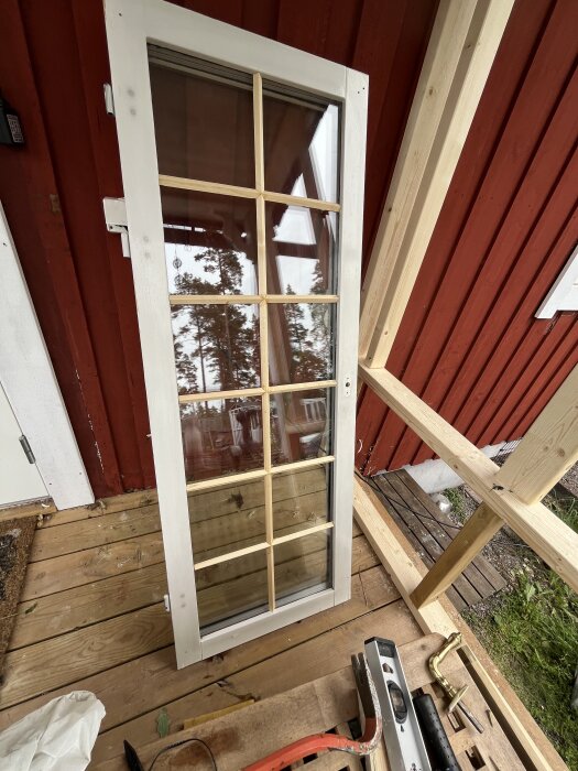 Fönster med galler väntar på montering, ståendes mot en röd trävägg på en veranda. Verktyg och mäthjälpmedel syns nedanför.